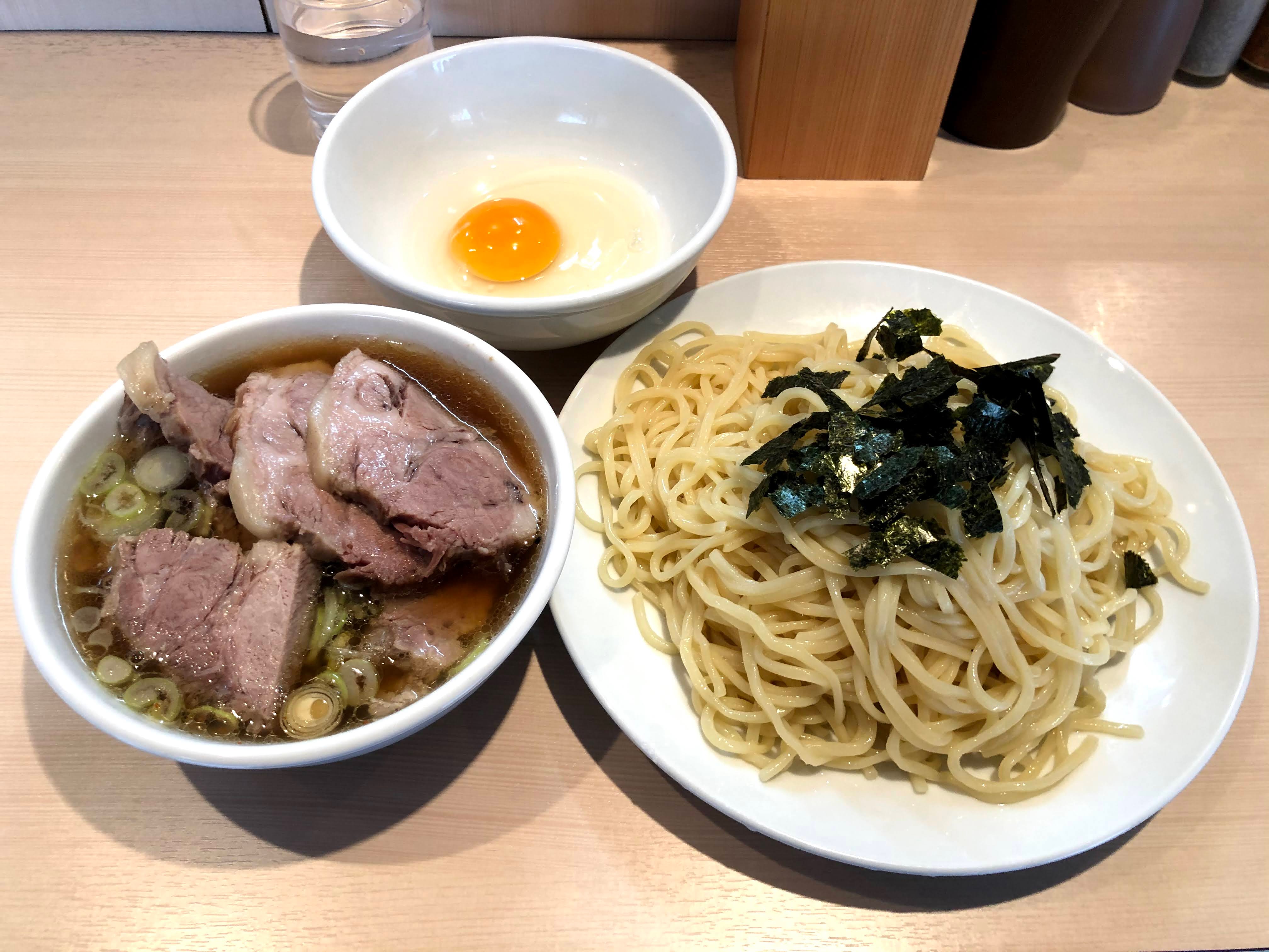 白っぽい木目調のカウンターに乗ったつけ麺。麺は中太で、白い皿に盛りつけれ、麺の上には揉み海苔がかけられている。つけ汁は澄んだ濃い茶色で、白い小鉢に入っている。つけ汁の小鉢には分厚いチャーシューがつけ汁から優にはみ出すほ山盛りになっており、輪切りの白ネギも浮かんでいる。更につけ汁と同じ大きさの小鉢に入った生玉子も添えられている。